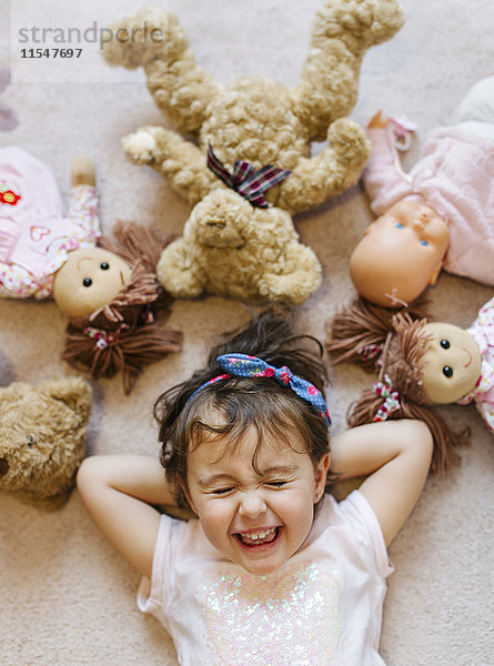 Porträt eines lachenden kleinen Mädchens auf dem Boden liegend mit Teddys und Puppen um sie herum.