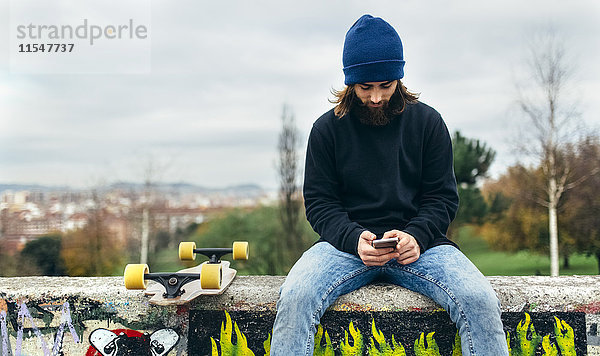 Ein bärtiger junger Skateboarder sitzt auf einer Wand und schaut auf sein Smartphone.