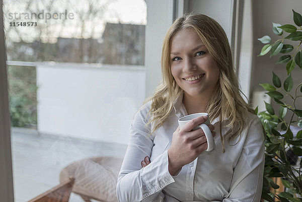 Porträt einer lächelnden jungen Frau mit einer Tasse Kaffee am Fenster