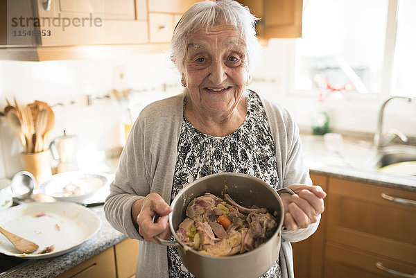 Porträt einer lächelnden Seniorin mit einem Kochtopf mit galicischem Eintopf in der Küche.