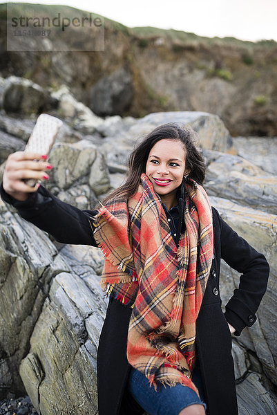 Spanien  Ferrol  Porträt einer lächelnden Frau  die einen Selfie mit ihrem Smartphone vor Felsen nimmt.