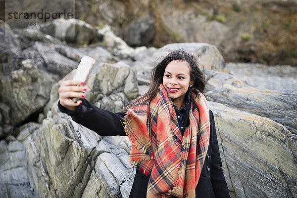 Spanien  Ferrol  Porträt einer lächelnden Frau  die einen Selfie mit ihrem Smartphone vor Felsen nimmt.