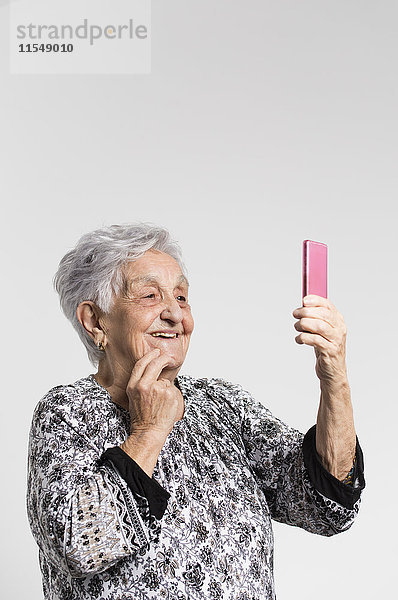 Porträt einer lächelnden Seniorin  die sich mit einem Smartphone vor weißem Hintergrund selbstständig macht.