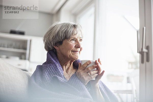 Porträt einer fröhlichen Seniorin auf der Couch zu Hause bei einer Tasse Kaffee