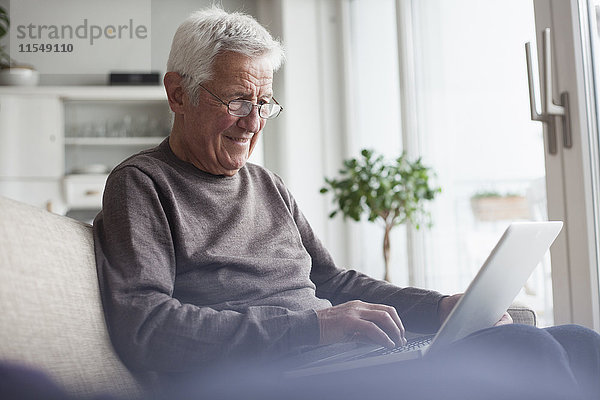 Porträt eines älteren Mannes  der zu Hause auf der Couch sitzt und einen Laptop benutzt.