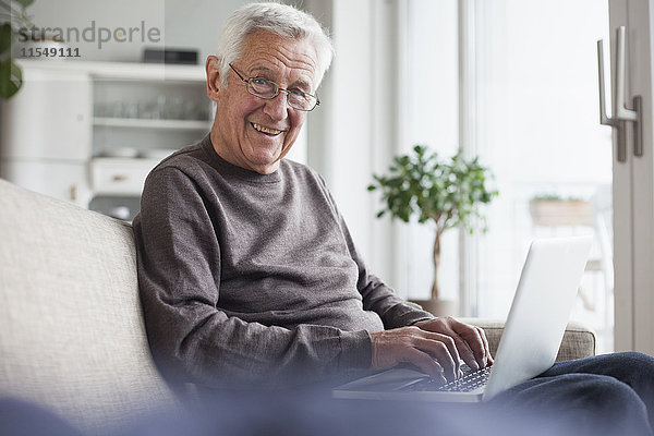 Porträt eines lächelnden älteren Mannes  der zu Hause auf der Couch sitzt und einen Laptop benutzt.