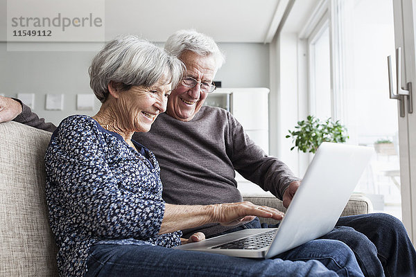 Glückliches Seniorenpaar sitzt auf der Couch im Wohnzimmer mit Laptop