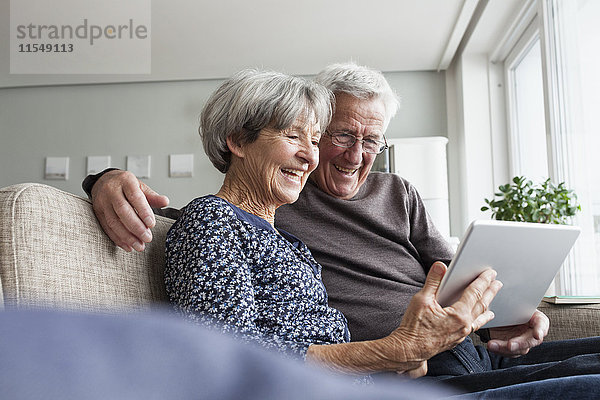 Glückliches Seniorenpaar sitzt auf der Couch im Wohnzimmer mit digitalem Tablett