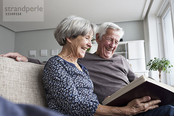 Lachendes Seniorenpaar sitzt auf der Couch im Wohnzimmer und schaut sich ein Fotoalbum an.