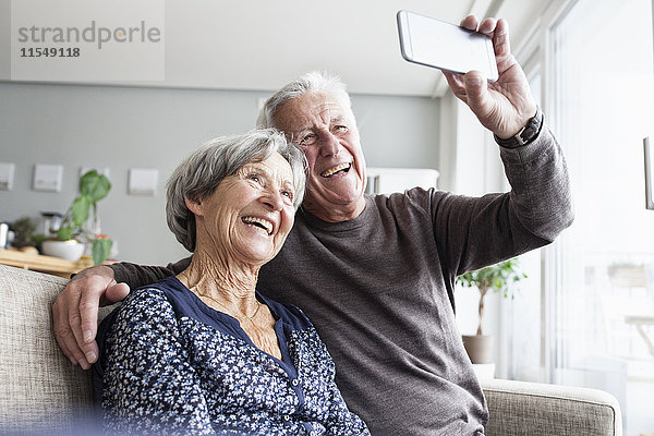 Lachendes älteres Paar  das auf der Couch im Wohnzimmer sitzt und Selfie mit Smartphone nimmt