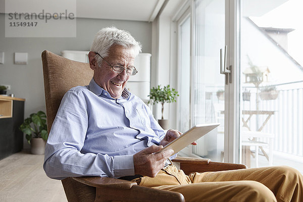 Porträt eines glücklichen älteren Mannes  der zu Hause auf einem Sessel sitzt und ein digitales Tablett benutzt.