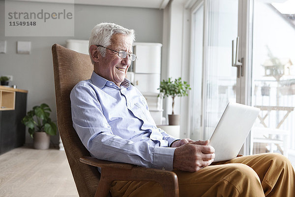 Porträt eines älteren Mannes  der zu Hause auf einem Sessel sitzt und einen Laptop benutzt.