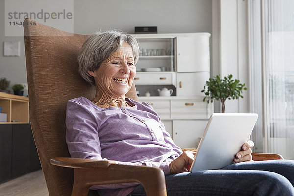 Porträt einer älteren Frau  die zu Hause auf einem Sessel sitzt und ein digitales Tablett benutzt.