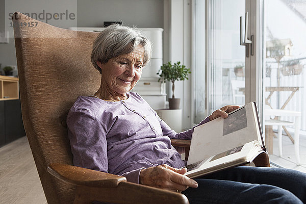 Porträt einer älteren Frau  die zu Hause auf einem Sessel sitzt und ein Fotoalbum anschaut.