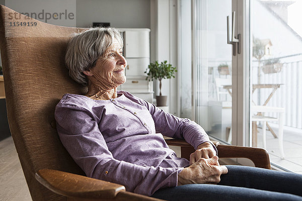 Porträt einer älteren Frau  die zu Hause auf einem Sessel sitzt und durchs Fenster schaut.