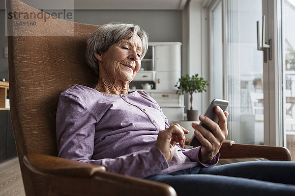 Porträt einer älteren Frau  die zu Hause auf einem Sessel sitzt und ein Smartphone benutzt.