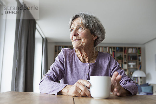 Porträt einer älteren Frau am Tisch mit einer Tasse Kaffee  die durchs Fenster schaut.