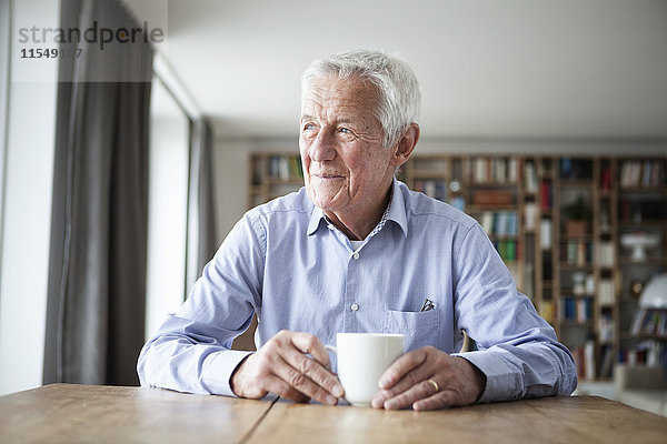 Porträt eines nachdenklichen älteren Mannes  der bei einer Tasse Kaffee am Tisch sitzt und durchs Fenster schaut.