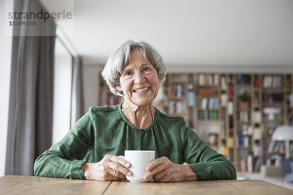 Porträt einer lächelnden Seniorin am Tisch mit einer Tasse Kaffee