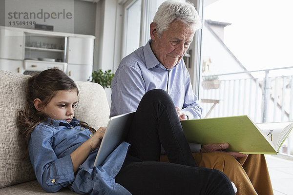 Großvater und seine Enkelin sitzen zusammen auf der Couch mit Buch und digitalem Tablett.