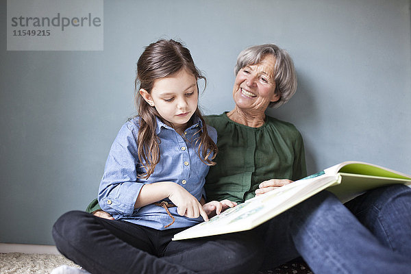 Glückliche Großmutter und ihre Enkelin sitzen mit einem Buch auf dem Boden.