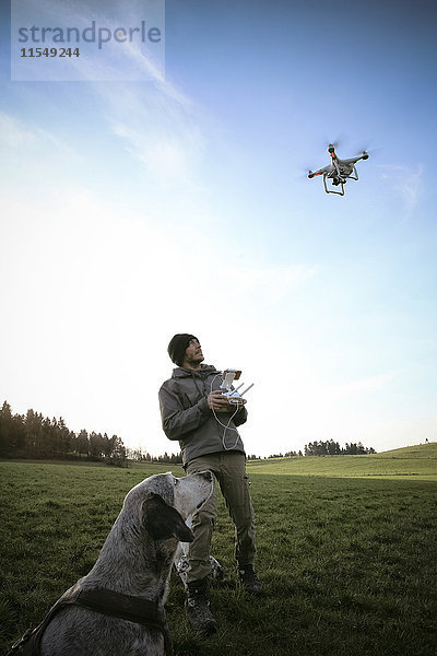 Mann auf einer Wiese fliegt Drohne  während sein Hund zusieht.