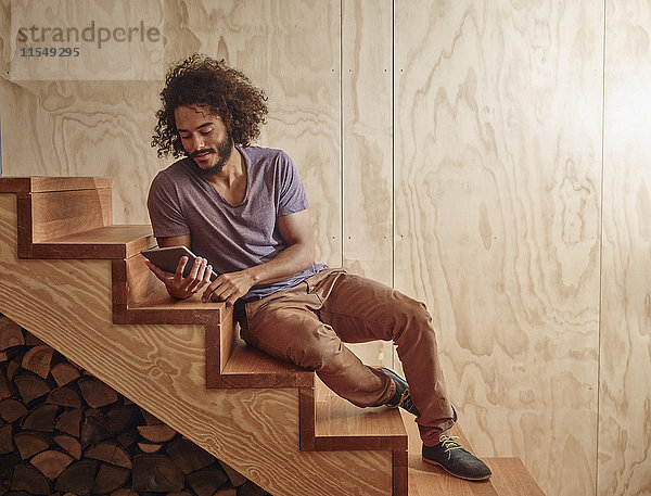 Junger Mann auf Holztreppe mit Blick auf digitales Tablett
