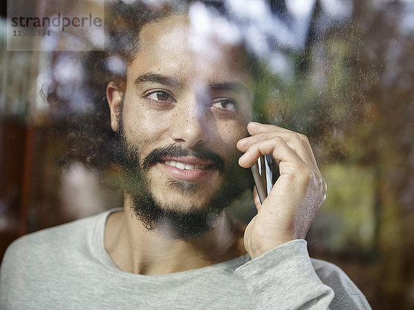 Lächelnder junger Mann auf dem Handy hinter der Fensterscheibe