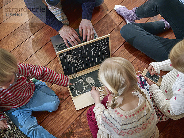Vater mit Laptop auf dem Boden sitzend mit Töchtern Zeichnung auf Spielzeug-Laptop
