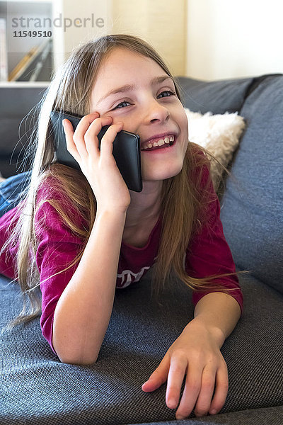 Porträt des lächelnden Mädchens auf der Couch liegend telefonierend mit Phablet