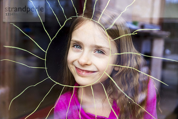 Porträt eines lächelnden Mädchens  das durch eine Fensterscheibe mit bemalter Sonne blickt.