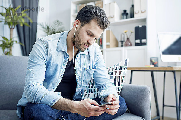 Junger Mann zu Hause mit Ohrhörern beim Blick auf das Smartphone