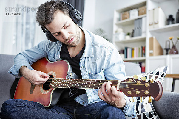 Junger Mann zu Hause auf der Couch sitzend  Kopfhörer tragend und Gitarre spielend