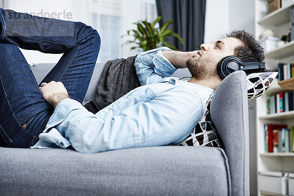 Junger Mann zu Hause auf der Couch liegend mit Kopfhörer