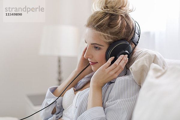 Porträt einer lächelnden blonden Frau  die auf dem Bett sitzt und Musik mit Kopfhörern hört.