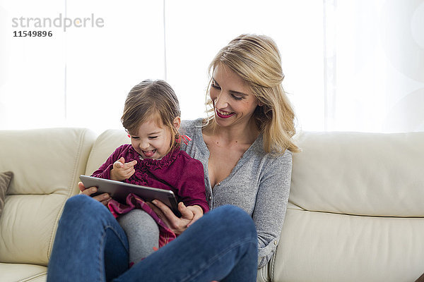 Mutter und ihre kleine Tochter sitzen auf der Couch im Wohnzimmer und schauen auf ein digitales Tablett.