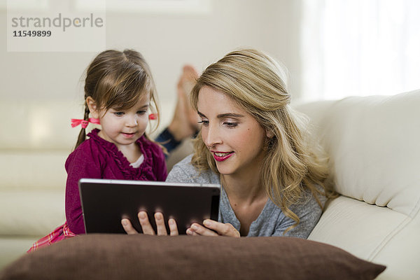 Mutter und ihre kleine Tochter zusammen auf der Couch im Wohnzimmer mit Blick auf das digitale Tablett