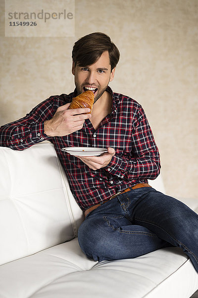 Porträt eines Mannes  der auf der Couch sitzt und ein Croissant isst.