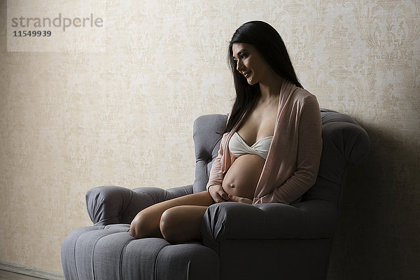 Lächelnde schwangere Frau in Dessous auf Sessel sitzend