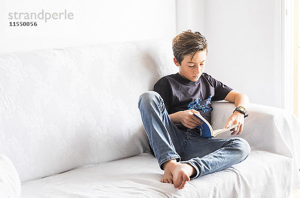 Junge sitzt auf einer weißen Couch und liest ein Buch.