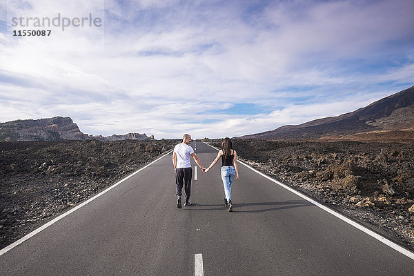 Spanien  Teneriffa  Rückansicht eines Paares  das Hand in Hand auf einer leeren Straße im Teide-Nationalpark geht.