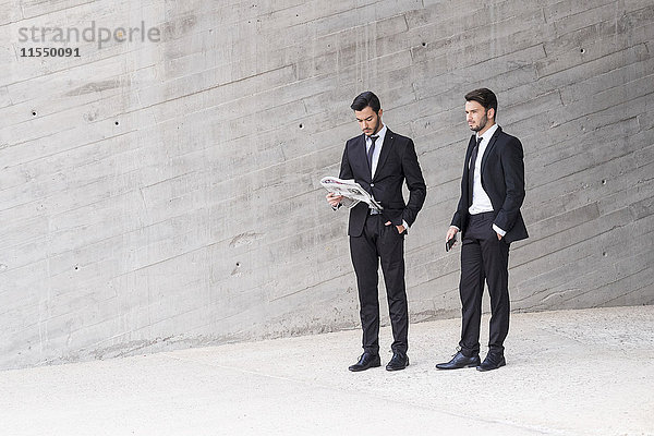 Zwei Geschäftsleute in schwarzen Anzügen stehen vor der Betonmauer