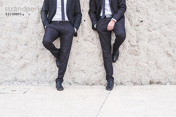 Zwei Geschäftsleute in schwarzen Anzügen lehnen sich nebeneinander an die Wand.