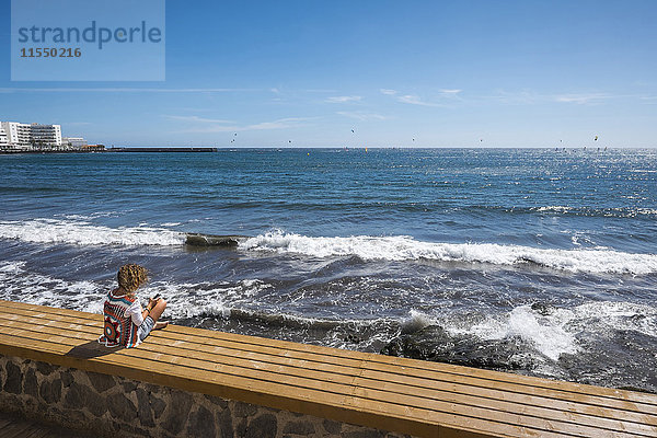 Spanien  Teneriffa  Frau auf einer Bank vor dem Meer sitzend mit ihrem Smartphone