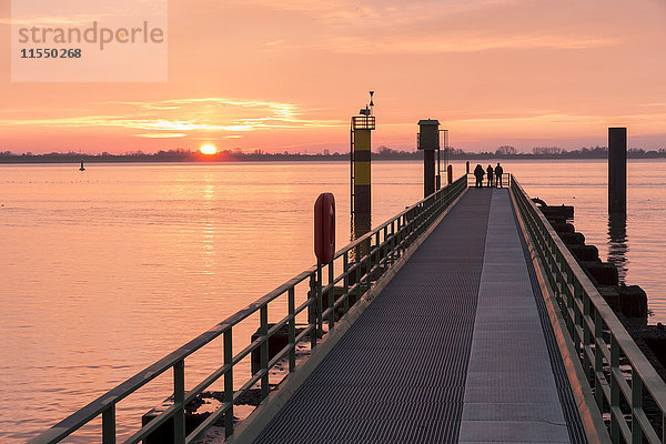 Deutschland  Bremerhaven  Silhouetten von drei Personen  die auf einem Steg stehen und den Sonnenuntergang beobachten.