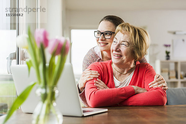 Lächelnde junge Frau umarmt Seniorin am Tisch mit Laptop