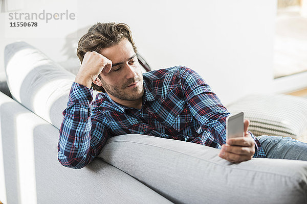 Porträt eines jungen Mannes  der auf der Couch sitzt und auf sein Smartphone schaut.
