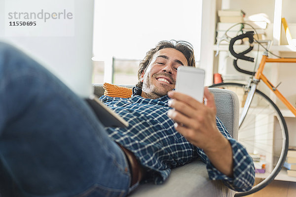 Junger Mann auf der Couch liegend mit Laptop auf sein Smartphone schauend