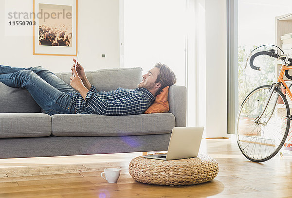Junger Mann zu Hause auf der Couch liegend mit Blick auf das digitale Tablett