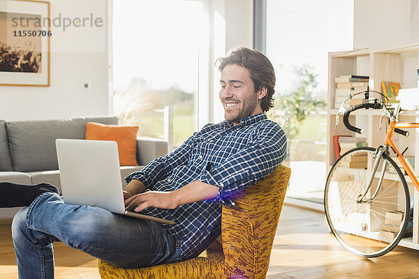 Porträt eines lächelnden jungen Mannes  der auf einem Sessel im Wohnzimmer mit einem Laptop sitzt.